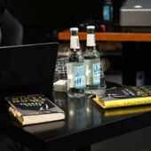 Zwei Salonfähig-Ausgaben und zwei Wasserflaschen vor einem Laptop auf einem schwarzen Tisch.
