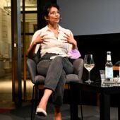 Elisa Aseva sits on a chair on an audience and speaks. She wears a clip-on microphone, and on a coffee table in front of her there is a glass of white wine.