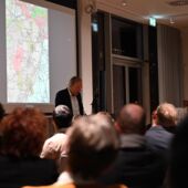 Michael Mönninger stands at the lectern and speaks to the audience. A map of Berlin-Wilmersdorf is projected behind him.