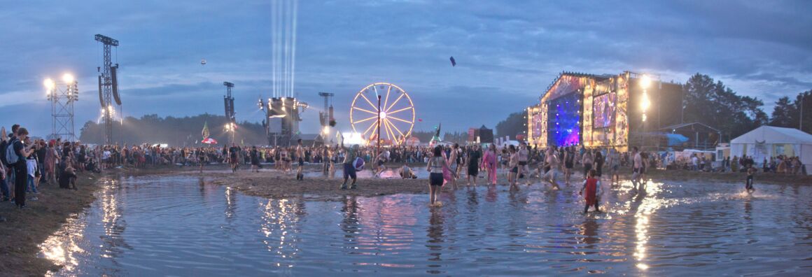 Scene from the Przystanek Woodstock festival. A large puddle can be seen in the evening light, with people dancing and playing. A brightly lit stage can be seen at the back right and a Ferris wheel in the middle.