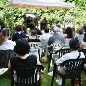 Im Garten des Literaturhauses sitzen circa 20 Personen auf Gartenstühlen, sie tragen Headsets und schauen auf ein Gartenzelt mit Bühne, auf der Ulrike Vedder mit Yoko Tawada spricht.